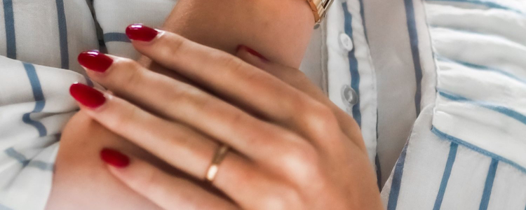 professional young woman with dark red nails holds her arm; she is wearing a gold watch and wedding band and wearing a white and blue striped button-frront blouse
