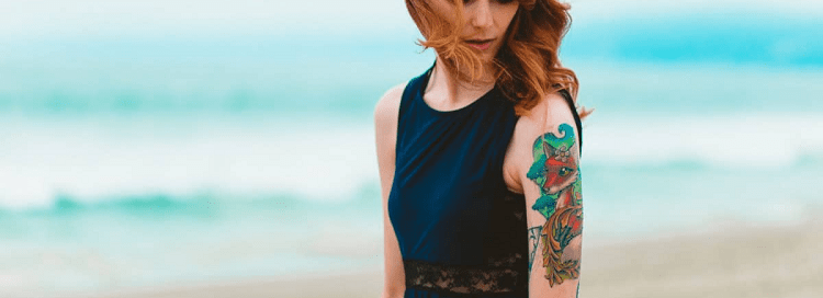 redhead on beach wears black tank with colorful tattooed arms