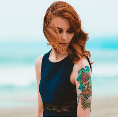 redhead on beach wears black tank with colorful tattooed arms