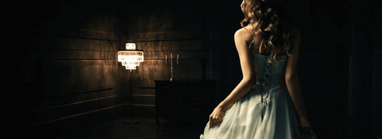 woman wears a ballgown in a dark room; she is turned away from the camera and moving toward a fancy standing light