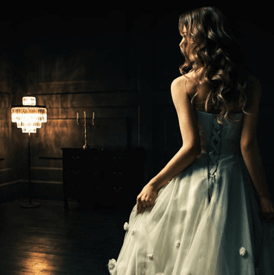 woman wears a ballgown in a dark room; she is turned away from the camera and moving toward a fancy standing light