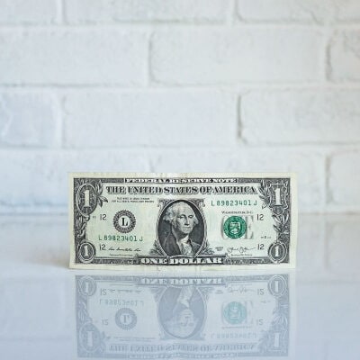 dollar bill sits on a shiny white counter; there is a matte white brick wall behind the dollar bill