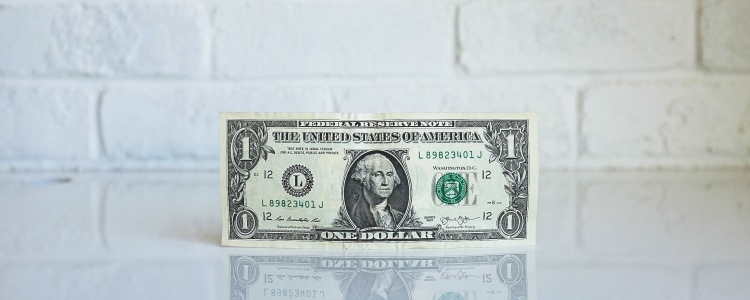 dollar bill sits on a shiny white counter; there is a matte white brick wall behind the dollar bill