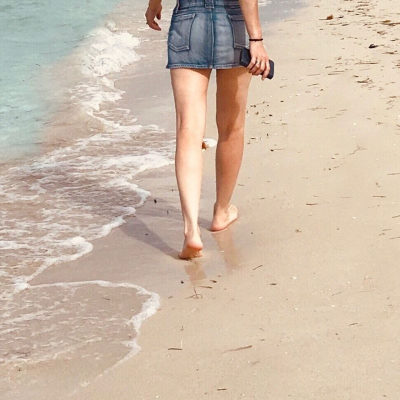 woman with hair-free legs walks along beach; she is wearing a denim miniskirt
