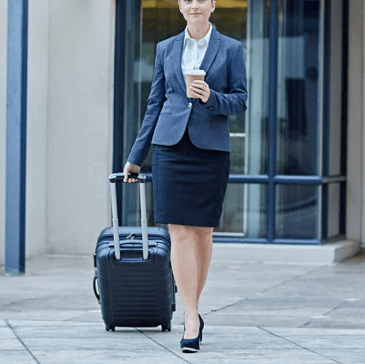 woman wearing skirt suit walking confidently outside; she is pulling a rolling bag behind her and carrying a cup of coffee.