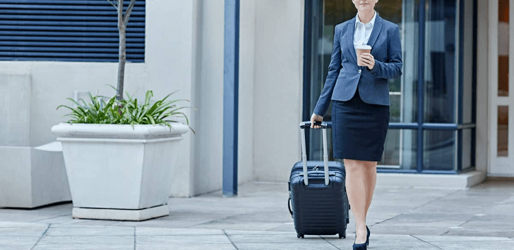 professional woman walking with a rolling back and a cup of coffee, wearing a blazer, skirt and white blouse