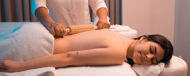 woman smiles blissfully while lying face down on a massage table; another person stands behind her rolling a pin on her bare back. 