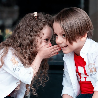 little girl whispers to a little boy; they are both wearing white tops
