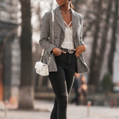 woman walks outside; she is wearing a plaid blazer, black ripped jeans, and a lacy top; she carries a white bag with pearl accents