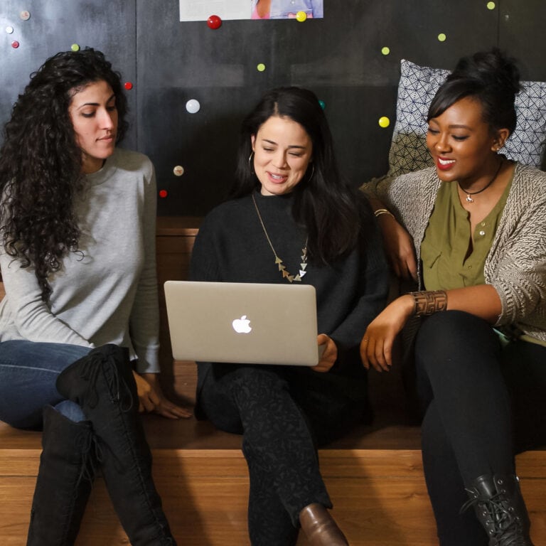 three stylish professional women look at a laptop; they're sitting on a brown bench and there is a black wall with gold dots behind them