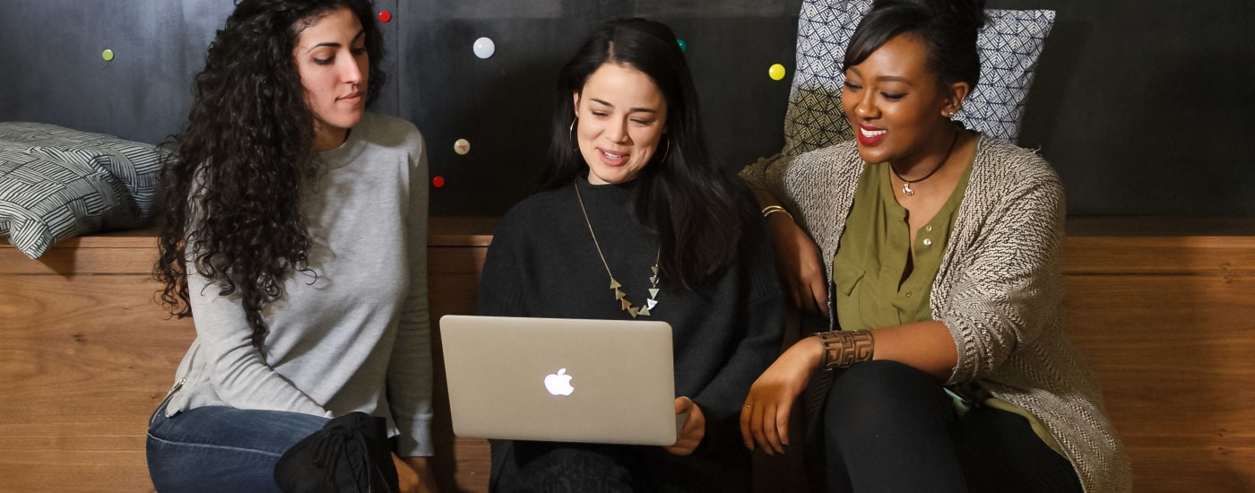 three stylish professional women look at a laptop; they're sitting on a brown bench and there is a black wall with gold dots behind them