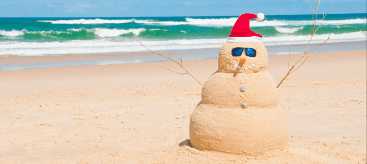 snowman made out of sand, on the beach, with a red and white Santa cap on