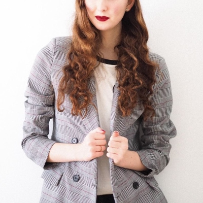 woman with red hair wears an oversized double-breasted blazer over a ringer tee; she is pulling the blazer closed and is wearing dark red lipstick