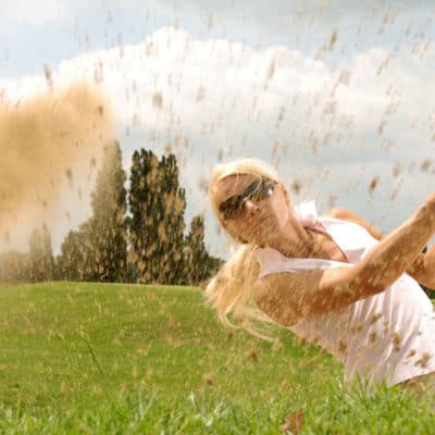 blonde woman swings golf club and sand sprays towards the camera; she might be attending a company golf outing
