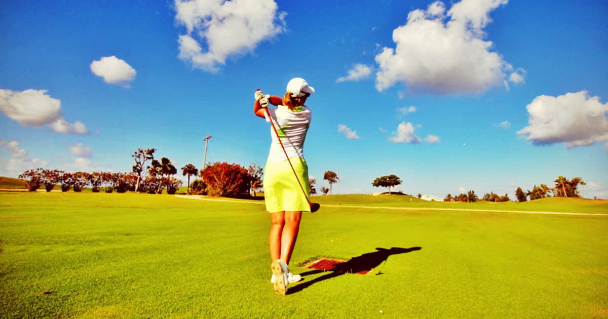 woman swings golf club on golf course; she is wearing a lime golf skort, a white polo, and a white hat