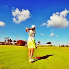 woman swings golf club on golf course; she is wearing a lime golf skort, a white polo, and a white hat