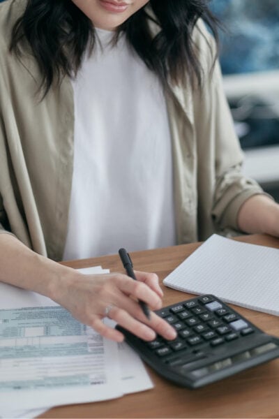 woman types on calculator; she has a notebook and spreadsheets open in front of her and is wearing a beige blazer and white tee