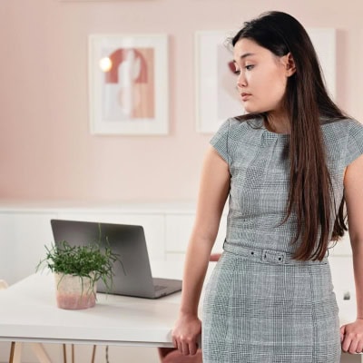 woman wears a houndstooth print dress; she has a sad look because she feels like she can't have MORE fun with patterns and colors with her corporate style -- she is in a pink office and has long black hair; there is a laptop and plant on the table behind the woman