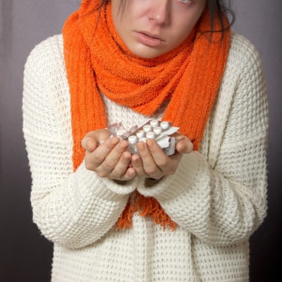 woman wears orange scarf and white sweater; her nose and eyes are read and she's holding handfuls of pills. 