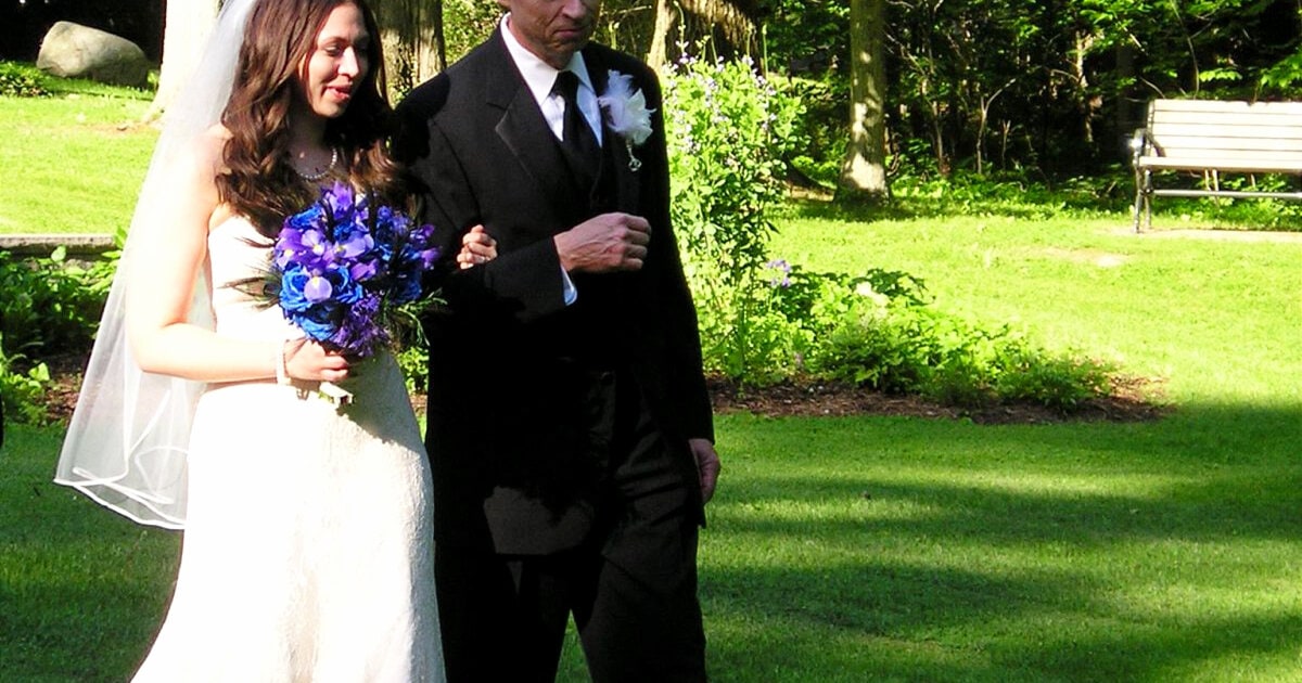 Kat being walked down the aisle by her father; she has a white chantilly lace dress on and carries a blue and purple wedding bouquet