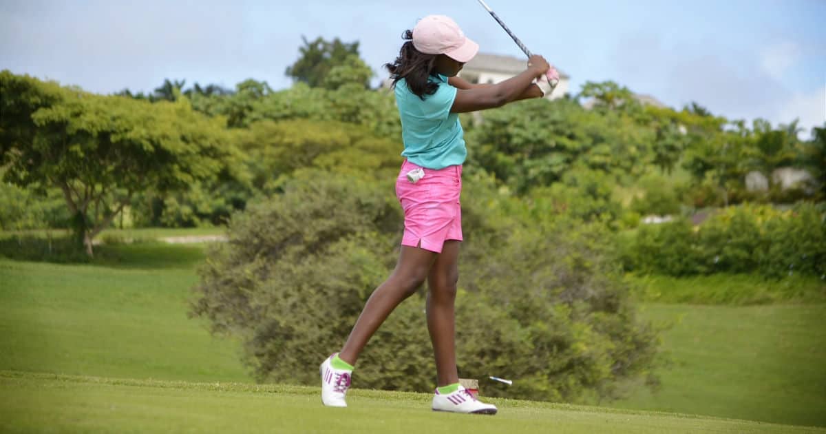 Black woman swings a golf club; she might be at a golf event for work! She is wearing pink shorts, a teal top, a white hat, and sneakers.