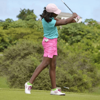 Black woman swings a golf club; she might be at a golf event for work! She is wearing pink shorts, a teal top, a white hat, and sneakers.