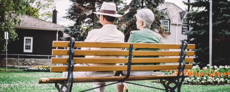 two elderly people sit on a bench; one wears a hat