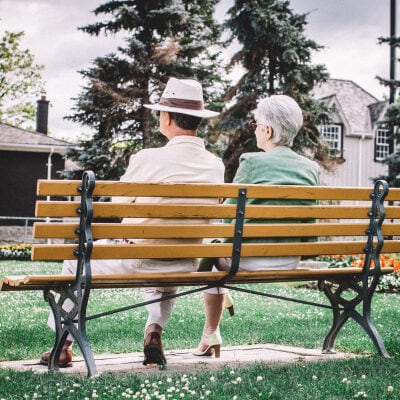 two elderly people sit on a bench; one wears a hat