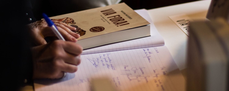 woman writes in a notebook; book next to her reads VIA FORA!