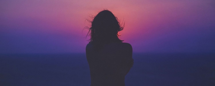woman holds her arms tightly; she stands in shadow in front of a very purple sunset/background