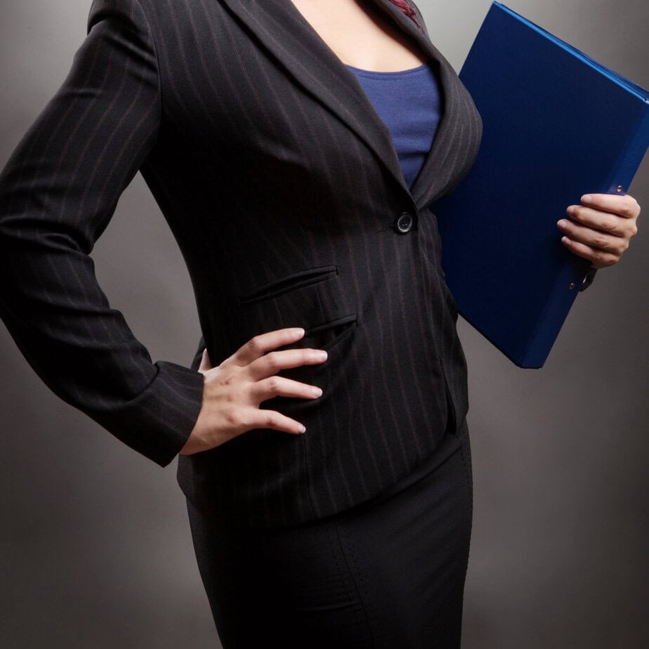 a professional woman holds a folder; she is wearing a blazer and does in fact look busty