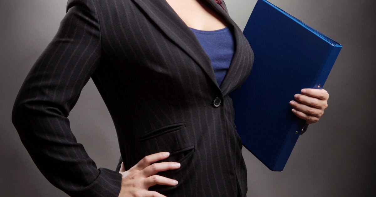 a professional woman holds a folder; she is wearing a blazer and does in fact look busty