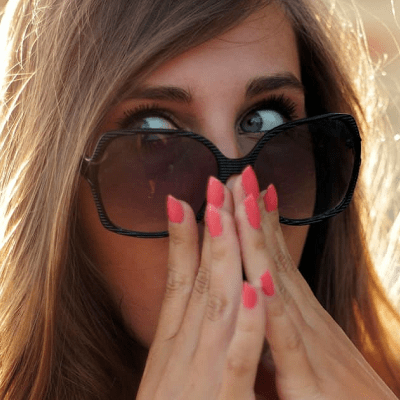 young woman wearing sunglasses has her hands to her nose; she looks surprised. She has pink fingernails and blonde hair.
