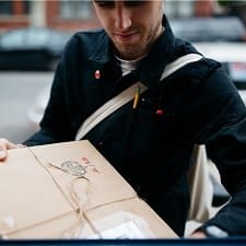 man wearing navy jacket with white crossbody is mailing a package wrapped in string.