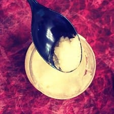 dark blue spoon being dipped into container of coconut oil; there is a purplish background