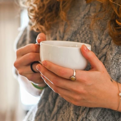 young woman holds a cup of tea; she is wearing rings and a gray sweater
