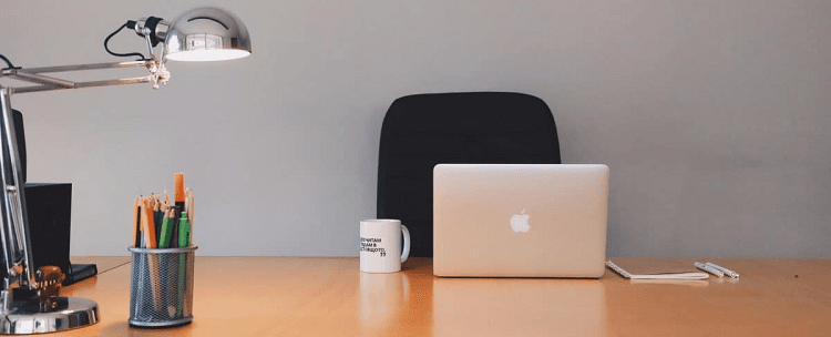 extremely clean desk area with a lamp, pencil cup, coffee mug, Apple laptop, notebook and pens on it. 