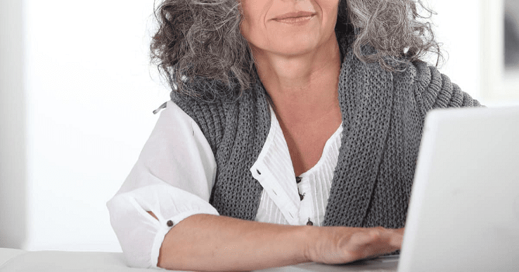 woman with gray hair works at a computer; she wears a crisp white blouse with a gray sweater over her shoulders