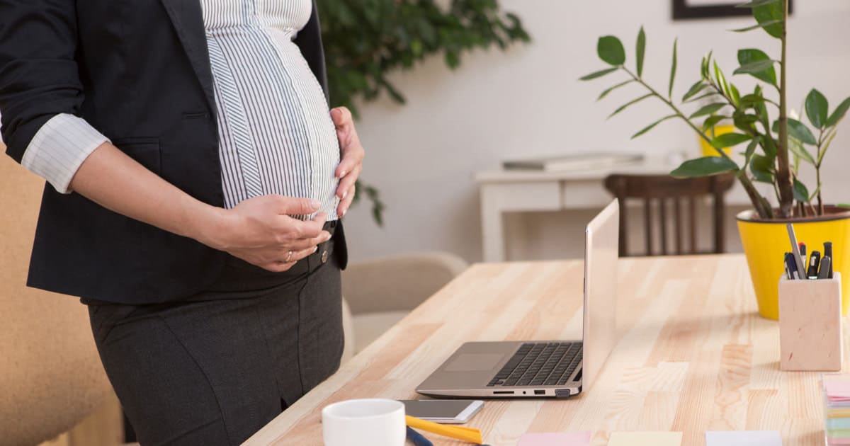 pregnant lawyer wearing a blazer, button-front blouse, and pencil skirt, standing in front of a laptop