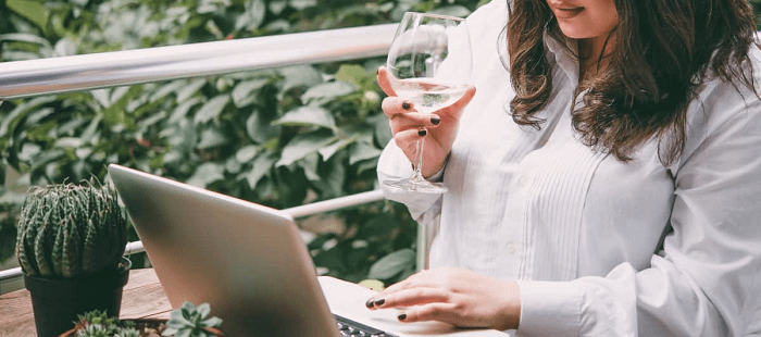 stylish professional woman wears plus-size white blouse, sips wine, and shops on her computer while sitting on a balcony