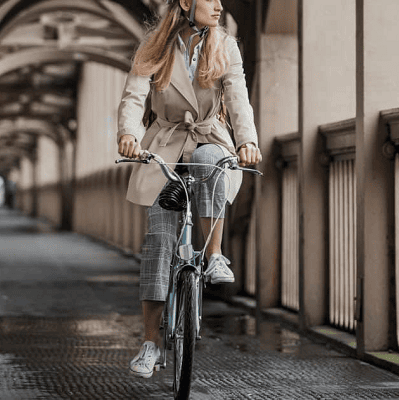 woman in beige trenchcoat wears helmet while she bikes to work through a covered corridor with beige arches, columns, and balcony railings