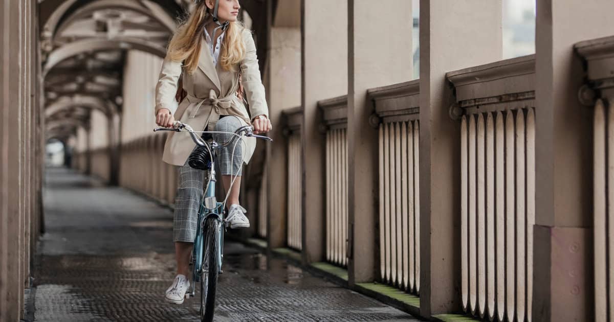 woman in beige trenchcoat wears helmet while she bikes to work through a covered corridor with beige arches, columns, and balcony railings