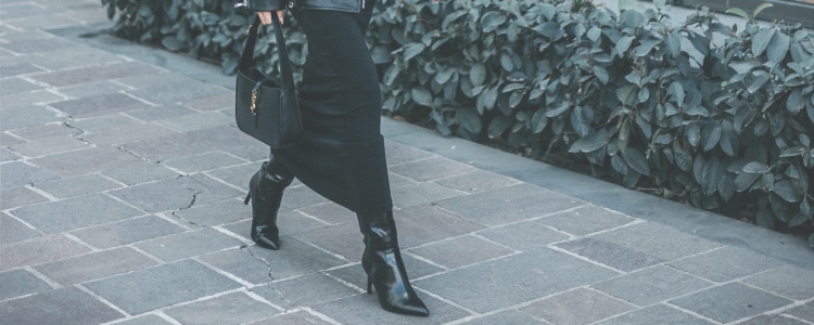 stylish professional woman walks down city street wearing stylish high-heeled boots for the office; the rest of her work outfit includes a midi dress, a handbag, and she's wearing a leather jacket for her commute