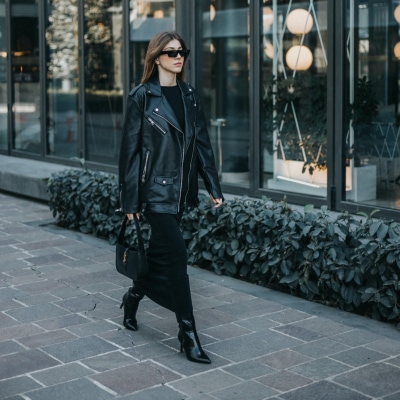 stylish professional woman walks down city street wearing stylish high-heeled boots for the office; the rest of her work outfit includes a midi dress, a handbag, and she's wearing a leather jacket for her commute