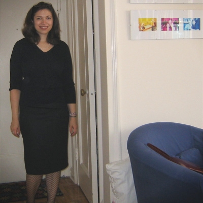 Kat in her apartment wearing navy sweater, navy skirt, and dark fishnet tights. 