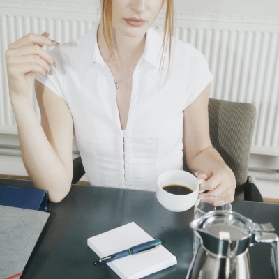 professional woman wears white top for spring with her work outfit; she is drinking an espresso and looking somewhat bored