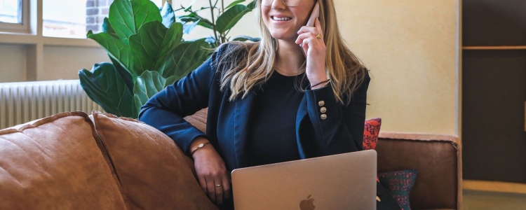 blond woman sits on brown leather couch with a laptop in her lap; her phone is to her ear. She wears a conservative office outfit of a navy blazer and black shirt. 