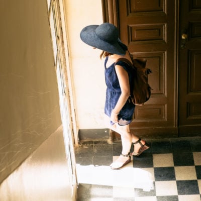 young woman exits a door; she is wearing a blue dress, a blue hat, and a backpack. The floor has a checkerboard pattern and the door is wooden