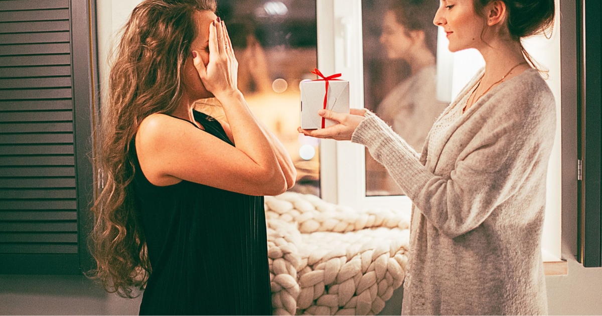 woman hiding eyes while second woman gives her a white box wrapped with a ribbon