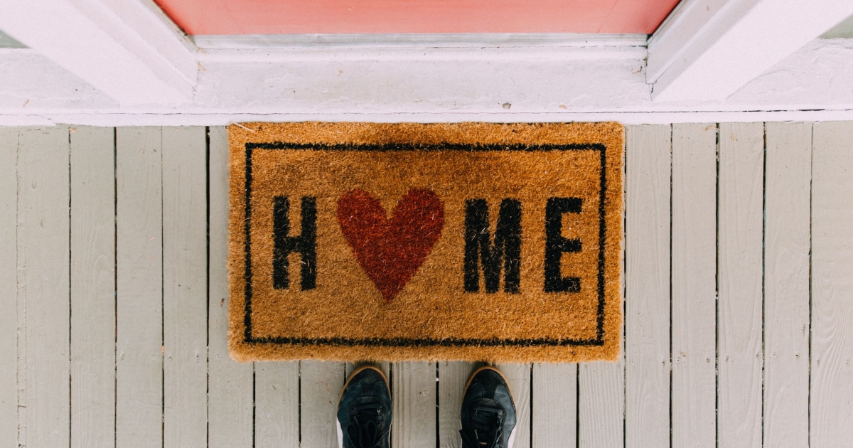 person in black sneakers standing in front of doormat that says HOME and a pink door